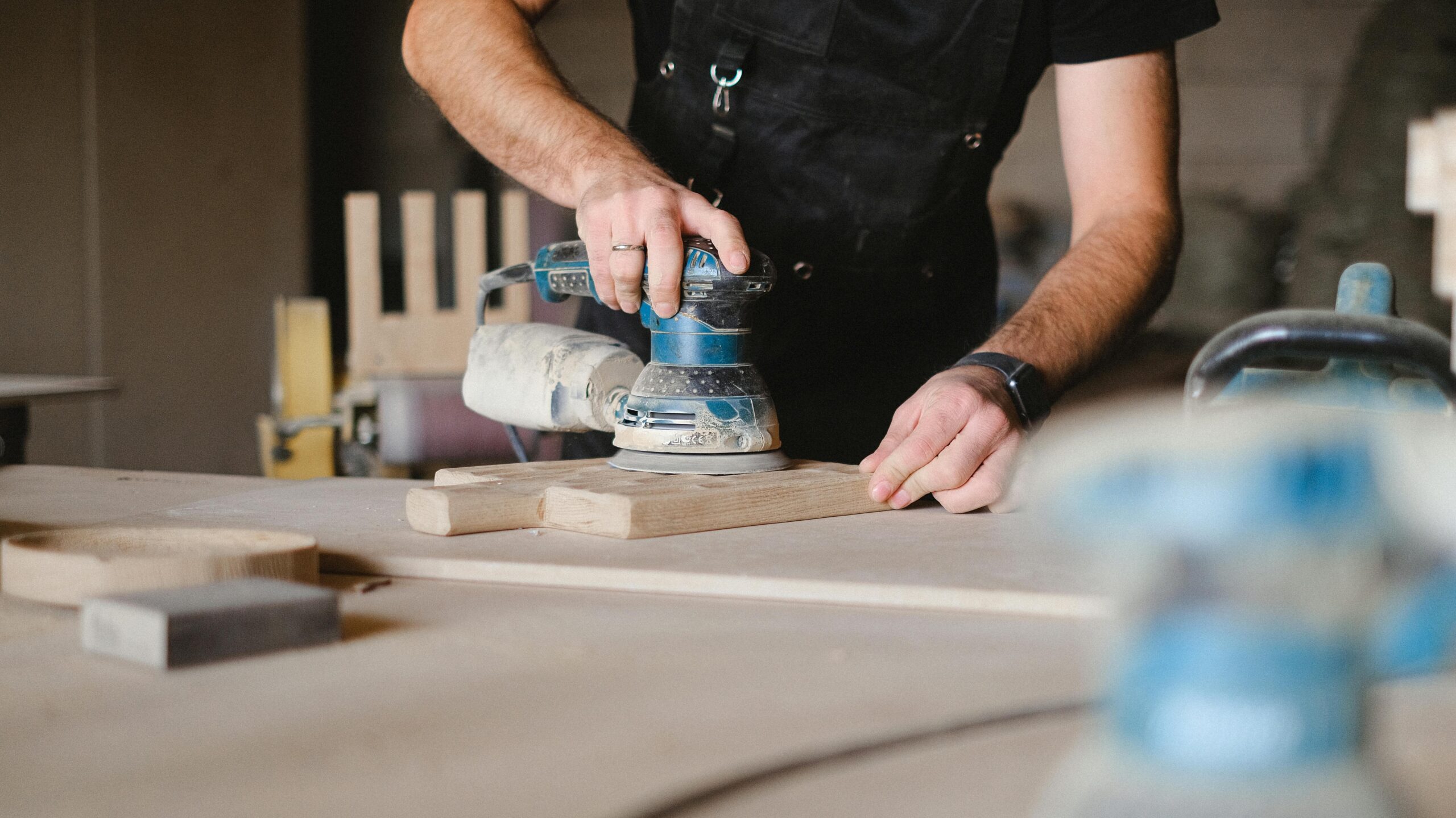 Unrecognizable male woodworker wearing uniform and polishing wooden board with random orbital sander at big table in professional studio